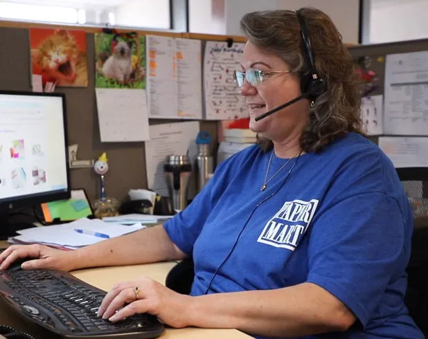 A woman wearing a blue 'Paper Mart' t-shirt and a headset sits at a desk in an office environment, typing on a keyboard.