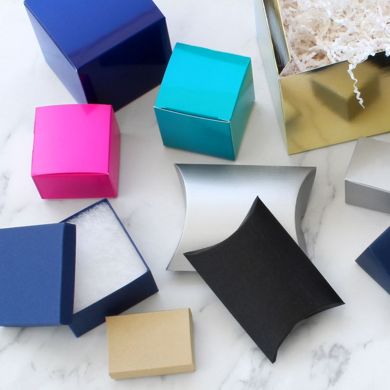 An assortment of colorful gift boxes and pillow boxes arranged on a white marble surface. The boxes come in various colors including navy blue, bright pink, teal, metallic gold, silver, black, and kraft beige, with some shown open to reveal white crinkle paper filler.